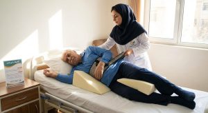 پرستار تیم کلینیک پاشنه در حال کمک به تغییر وضعیت بدن بیمار سالمند در بستر با استفاده از تجهیزات کمکی در منزل در تهران، به عنوان بخشی از برنامه مراقبتی و درمان زخم بسترهای عمیق.
