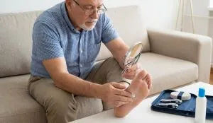 مرد سالمند دیابتی در حال معاینه دقیق کف پای خود با ذره‌بین برای پیشگیری از عفونت زخم در منزل.